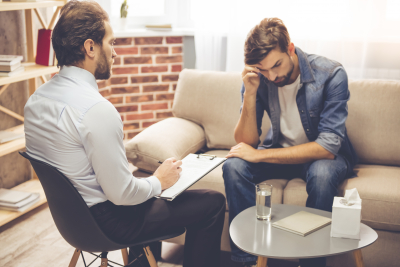 young man is sitting on couch and talking to the psychologist while doctor is making notes